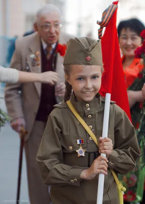 Фото Девочка в военной форме несет флаг СССР с георгиевской ленточкой, на  заднем плане идут ветераны ВОВ