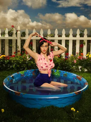 Фото Американская певица и актриса Кэти Перри / Katy Perry сидит в бассейне  на фоне цветов у белого забора и облаков в небе