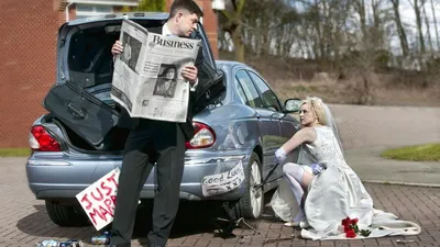 Обои на рабочий стол Невеста в белом свадебном платье меняет пробитое  колесо автомобиля, а жених невозмутимо читает газету, облокотившись на  открытый багажник, обои для рабочего стола, скачать обои, обои бесплатно