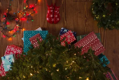 Обои на рабочий стол Ножки в красных новогодних носочках со снежинками  перед наряженной елкой с подарками под ней, рядом лежит венок и светящаяся  разноцветными огоньками гирлянда, обои для рабочего стола, скачать обои,